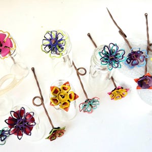 Puede incluir: Una colecci&oacute;n de objetos decorativos con forma de flor, coloridos y hechos a mano. Cada flor presenta dise&ntilde;os intrincados y tonos vibrantes, incluyendo rosa, amarillo, azul y morado. Est&aacute;n unidos a finos tallos marrones con un lazo en la parte superior.