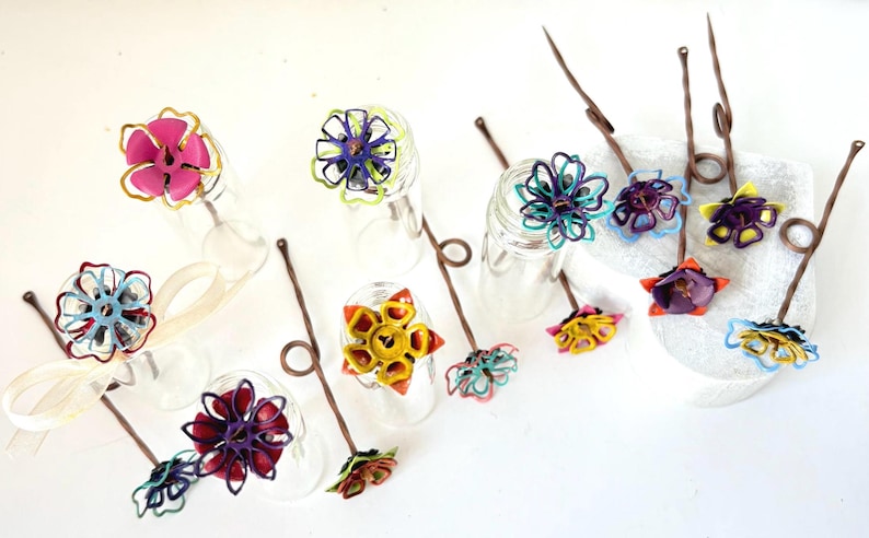 Puede incluir: Una colecci&oacute;n de objetos decorativos con forma de flor, coloridos y hechos a mano. Cada flor presenta dise&ntilde;os intrincados y tonos vibrantes, incluyendo rosa, amarillo, azul y morado. Est&aacute;n unidos a finos tallos marrones con un lazo en la parte superior.