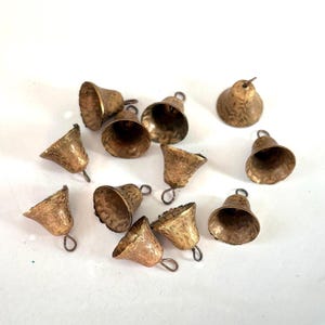 Könnte beinhalten: Eine Sammlung kleiner, strukturierter Messingglocken mit gehämmertem Finish. Jede Glocke hat eine Schlaufe zum Aufhängen. Die Glocken variieren in Größe und Form, verstreut auf einer weißen Oberfläche. Ideal für Bastelarbeiten oder Dekorationszwecke.