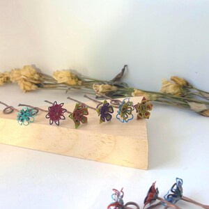Puede incluir: Colecci&oacute;n de seis horquillas para el cabello hechas a mano con forma de flor. Cada horquilla presenta una combinaci&oacute;n de colores &uacute;nica, incluyendo turquesa, burdeos, rojo, morado, amarillo y azul. Las horquillas est&aacute;n dispuestas sobre un bloque de madera, con flores secas en el fondo.