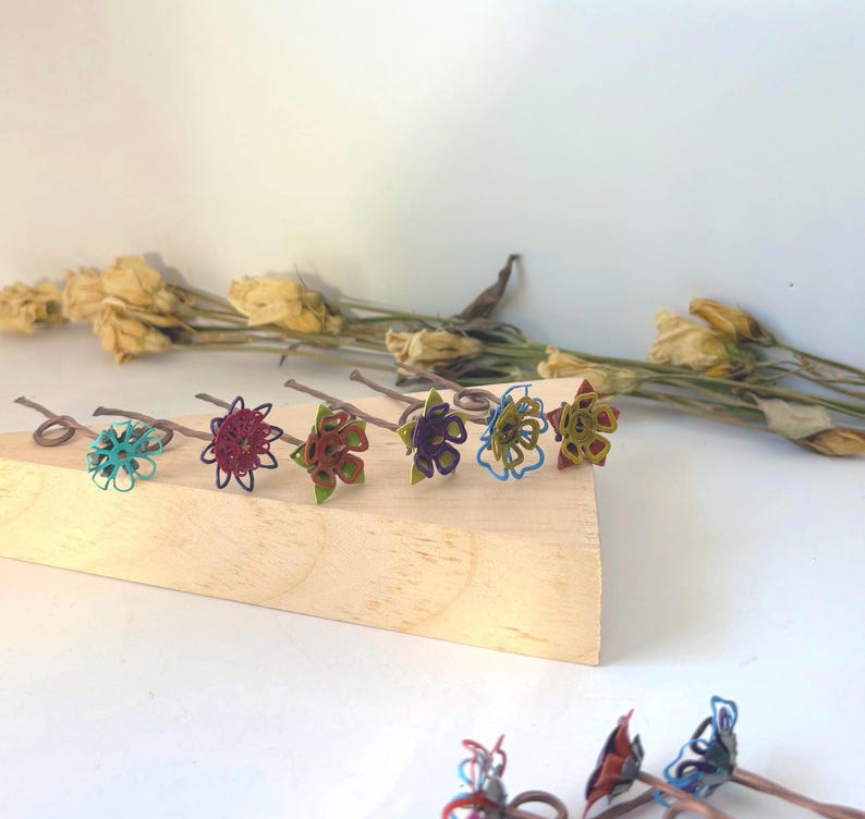 Puede incluir: Colecci&oacute;n de seis horquillas para el cabello hechas a mano con forma de flor. Cada horquilla presenta una combinaci&oacute;n de colores &uacute;nica, incluyendo turquesa, burdeos, rojo, morado, amarillo y azul. Las horquillas est&aacute;n dispuestas sobre un bloque de madera, con flores secas en el fondo.