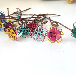 Puede incluir: Una colecci&oacute;n de horquillas para el pelo con flores hechas a mano. Cada horquilla presenta una flor de alambre en tonos rosa, amarillo, azul y verde, con tallos marrones. Las flores est&aacute;n dispuestas sobre una superficie blanca.