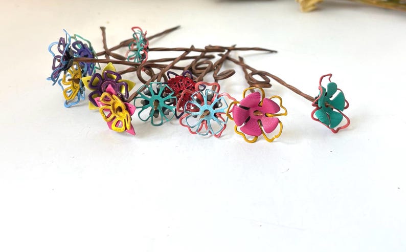 Puede incluir: Una colecci&oacute;n de horquillas para el pelo con flores hechas a mano. Cada horquilla presenta una flor de alambre en tonos rosa, amarillo, azul y verde, con tallos marrones. Las flores est&aacute;n dispuestas sobre una superficie blanca.
