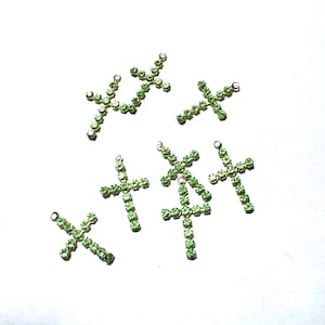 Puede incluir: Varios dijes de cruz dorados, cada uno adornado con filas de pequeños brillantes verdes claros. Las cruces están dispersas sobre una superficie blanca, con un pequeño lazo en la parte superior para sujetarlas a joyas. La estética general es delicada y brillante.