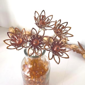 May include: A bouquet of copper-colored metal flowers in a clear glass vase. The flowers have intricate, openwork petals and a central bead. The vase is filled with small, golden-colored decorative elements. The background is white.