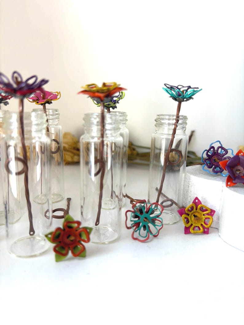 Puede incluir: Botellas de vidrio transparente con flores de alambre de colores y tallos marrones. Las flores son de varios colores, incluyendo rojo, amarillo, rosa y turquesa. Algunas flores est&aacute;n dentro de las botellas.