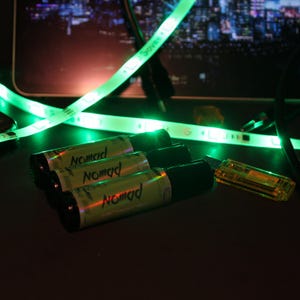 May include: A close-up shot of several cylindrical containers labeled "Nomad" with a black cap, alongside a small, clear, rectangular device. Green LED strip lights are visible in the background.