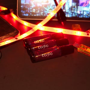 May include: Three dark red roll-on bottles with the word "corpo" printed on them. The bottles are arranged on a dark surface, with a red LED light strip and a USB drive in the background.