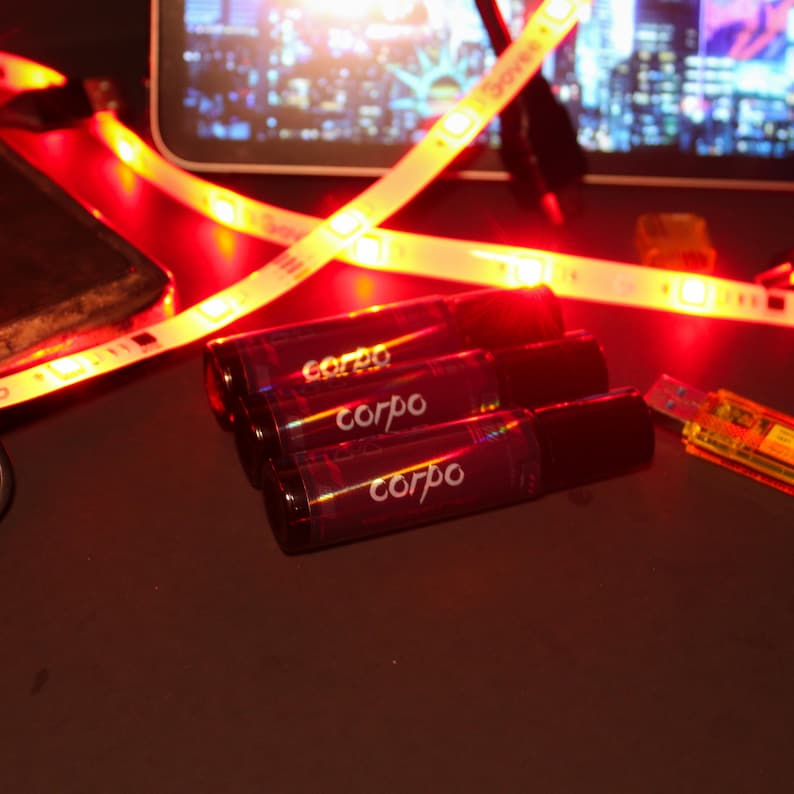 May include: Three dark red roll-on bottles with the word "corpo" printed on them. The bottles are arranged on a dark surface, with a red LED light strip and a USB drive in the background.