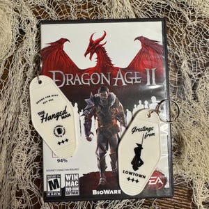 May include: A Dragon Age II video game case with two vintage-style keychains. One keychain reads "The Hanged Man" and the other says "Greetings from Lowtown". The game case features a red dragon and a warrior.