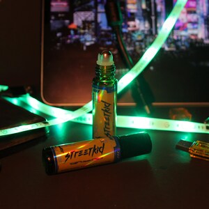 May include: Two roll-on bottles with the text "Streetkid" in a yellow and black design. The bottles are green and have a silver rollerball top. A green LED light strip and a cityscape background are visible.