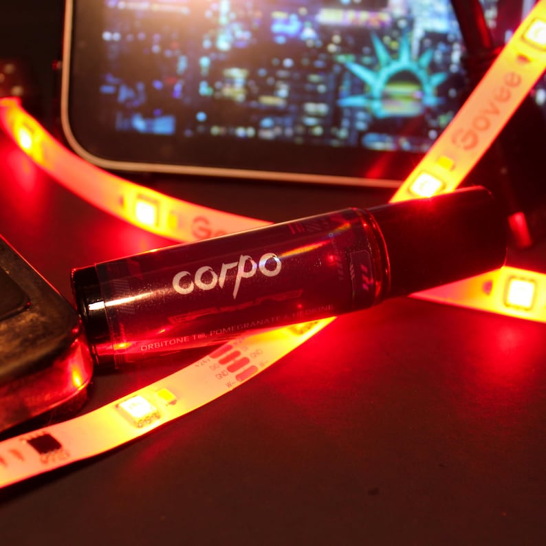 May include: A dark red glass bottle with the word "corpo" in white script. The bottle is lying on a surface with red and yellow LED strip lights. The background shows a blurred image of a city at night.