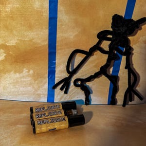 May include: Three small amber glass roller bottles with black caps, each featuring a tan label with the word REPLICATE printed in bold black capital letters. The bottles are arranged in a horizontal stack on a tan surface.