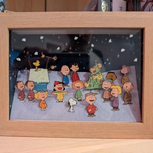 Puede incluir: Una caja de sombra enmarcada que presenta una escena invernal con personajes de dibujos animados. La escena incluye un árbol de Navidad decorado, nieve y varias figuras con trajes coloridos. El marco es de madera clara.