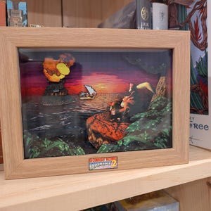 May include: Framed artwork depicting a sunset scene with a pixelated boat and characters. The artwork is in a light brown wooden frame. The image has a colourful sunset with a volcano and characters from the Donkey Kong Country 2 video game.