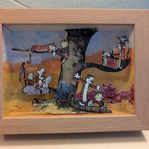 May include: A framed shadow box with a cartoon scene. The artwork shows a tree with cartoon characters, including a tiger and a boy, in different poses. The frame is made of light-coloured wood, and the background has a warm, autumnal colour palette.