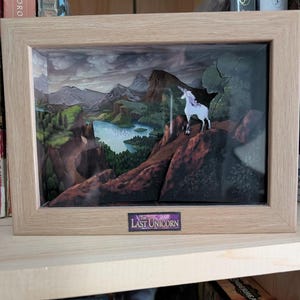 May include: A framed shadow box depicting a mountainous landscape with a lake and a white unicorn. The artwork is set in a wooden frame, with the title "Last Unicorn" at the bottom. The scene is a fantasy-themed art piece.