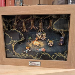 Puede incluir: Un diorama enmarcado que presenta una escena de pixel art de una fogata en un bosque. Varios personajes se reúnen alrededor del fuego. El marco es de madera clara y el diorama está sobre un fondo oscuro.