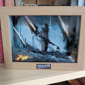 May include: Framed artwork featuring a figure in futuristic armor holding a weapon, set against a sci-fi backdrop with fiery elements. The artwork is in a light-colored wooden frame, with the "Mass Effect" logo at the bottom.