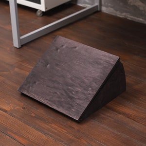 Puede incluir: Un reposapiés de madera teñida oscura con un diseño inclinado. El reposapiés está hecho de varias piezas de madera y se coloca sobre un suelo de madera. El reposapiés mide aproximadamente 30 cm de largo y 25 cm de ancho.