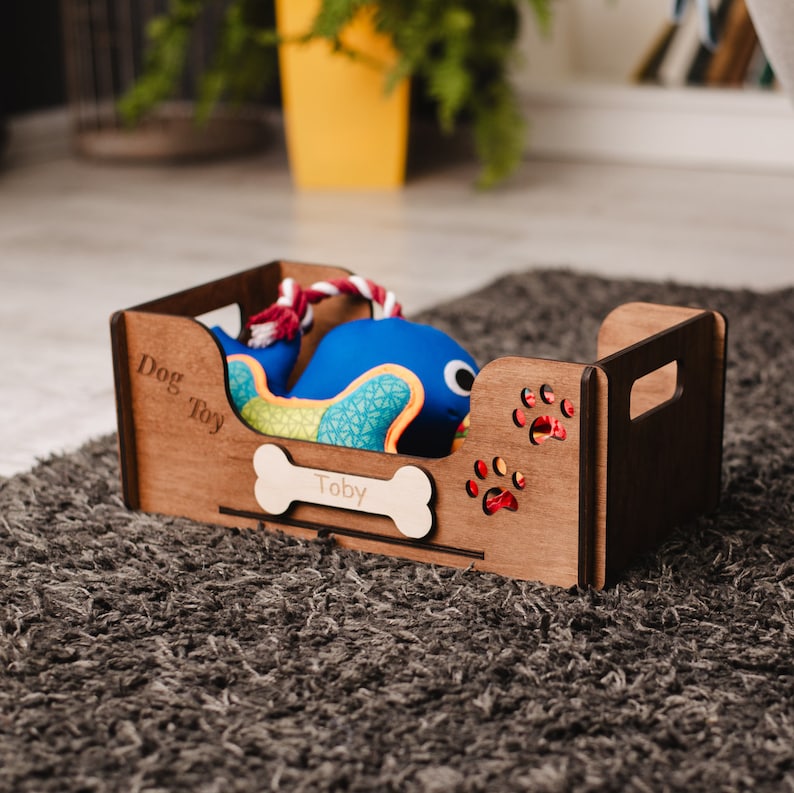 May include: A wooden dog toy storage box with a blue and green whale toy inside. The box has the words "Dog Toy" and "Toby" engraved on it. The box has paw print cutouts on the sides.