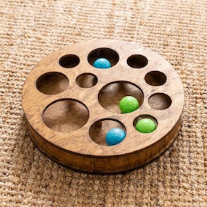 May include: A circular wooden puzzle board featuring various sized cutouts and holes. The natural brown wood grain is visible throughout the structure. Four small, smooth spheres in bright blue and lime green colors are placed within the openings.