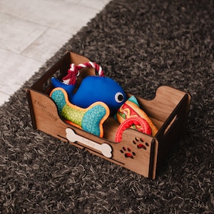 May include: A wooden dog toy box with the name "Toby" engraved on it. The box contains a blue whale plush toy, a colorful patterned toy, a red rope toy, and a hot dog shaped toy.
