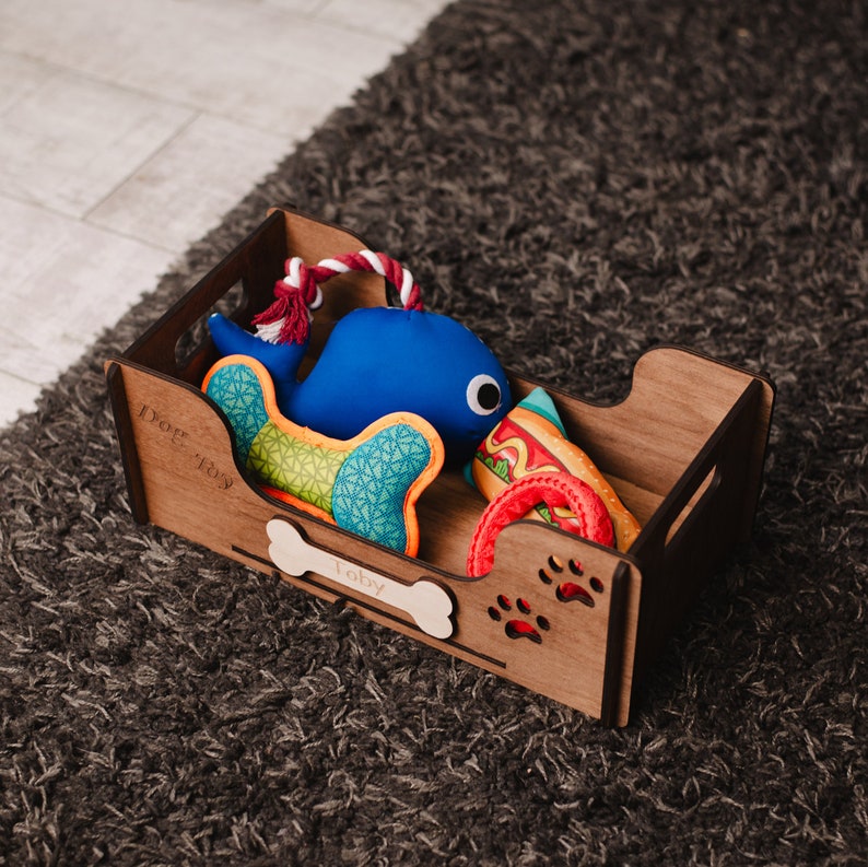 May include: A wooden dog toy box with the name "Toby" engraved on it. The box contains a blue whale plush toy, a colorful patterned toy, a red rope toy, and a hot dog shaped toy.