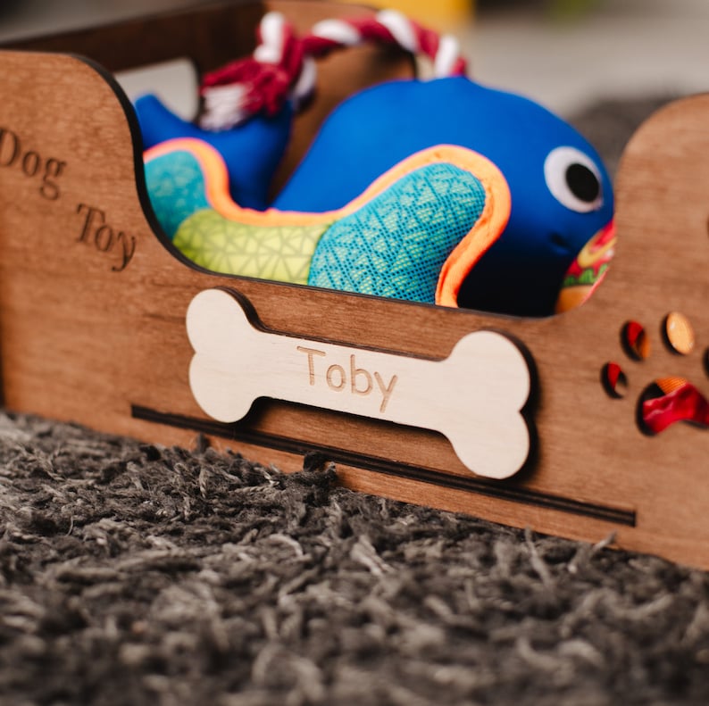 May include: A wooden dog toy box with a bone-shaped name tag that says "Toby". The box is filled with a blue and green dog toy.