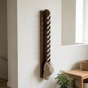 May include: A vertical dark brown wooden wall rack featuring ten evenly spaced hooks along its length. A plain beige baseball cap hangs from the bottom hook of the rack, which is mounted on a smooth white wall surface.