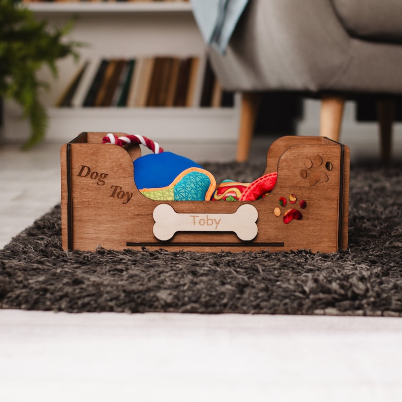 May include: A wooden dog toy storage box with the words "Dog Toy" and "Toby" engraved on it. The box is filled with various dog toys, including a blue squeaky toy, a red rope toy, and a green and yellow plush toy.