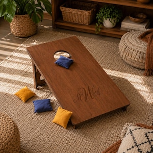 Könnte beinhalten: Ein rechteckiges Cornhole-Spielbrett aus Holz mit einem kreisförmigen Loch oben und einer individuellen Monogramm-Gravur unten. Das Set enthält vier quadratische Sitzsäcke in den Farben Blau und Gelb, die auf und um das Brett liegen.