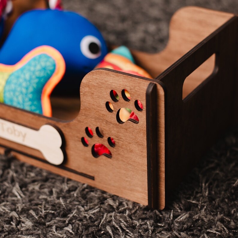 May include: A wooden dog toy storage box with the name "Toby" engraved on the front. The box has paw print cutouts and is filled with colorful plush toys.