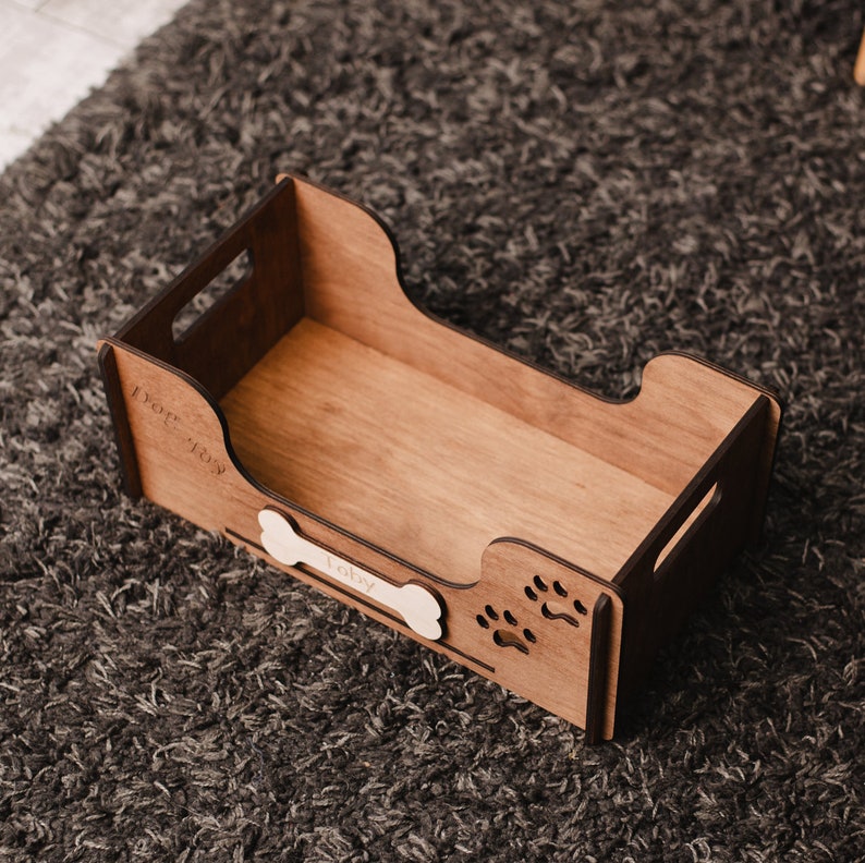 May include: A wooden dog bed with a white bone-shaped name tag that says "Toby". The bed has paw print cutouts on the side.