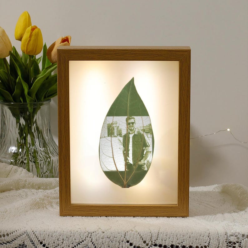 May include: A framed photo of a man in a leaf, illuminated by a light behind the frame. The frame is made of wood and has a light brown finish.