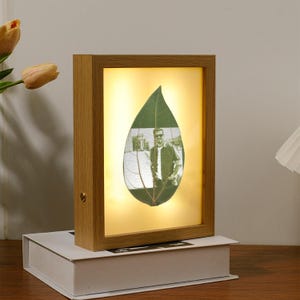 May include: A wooden picture frame with a light inside, showcasing a pressed leaf with a black and white photo of a person inside. The frame has a light brown finish.