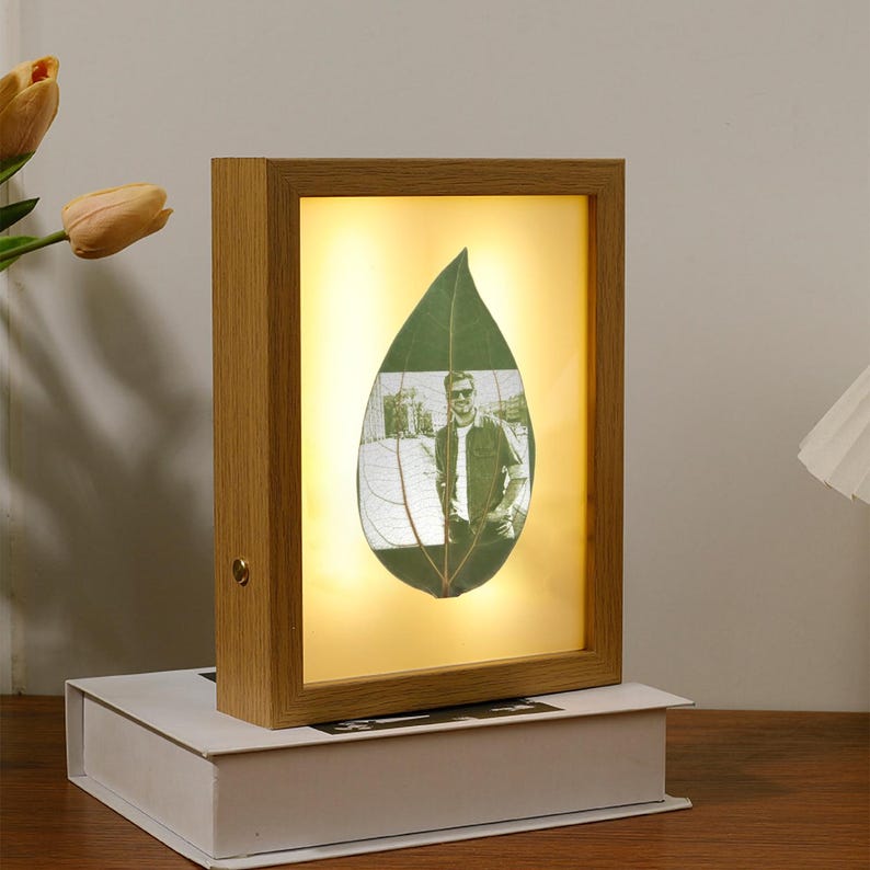 May include: A wooden picture frame with a light inside, showcasing a pressed leaf with a black and white photo of a person inside. The frame has a light brown finish.