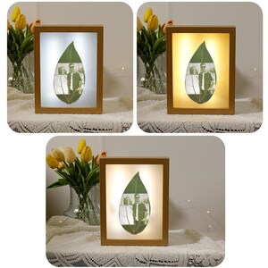 May include: A wooden frame with a light inside, showcasing a pressed leaf with a photo of a person inside. The frame is on a table with a vase of yellow flowers.