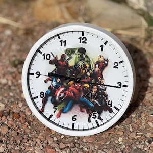 May include: White round clock with black hands and black numbers. The clock face features a graphic of various Marvel superheroes, including Spider-Man, Captain America, Hulk, Wolverine, Iron Man, and Thor.