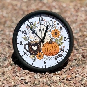 May include: A round wall clock with a black frame and a white face featuring a graphic illustration of a ghost, an orange pumpkin, and a dark mug with a heart design, surrounded by autumn-themed floral elements and black clock hands.