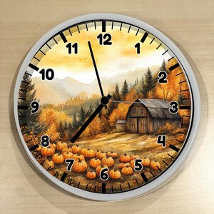May include: A round wall clock with a white frame featuring a watercolor illustration of a rustic barn in an autumn landscape with orange trees and a field of pumpkins. The clock face has black numbers from 1 to 12 and black hour and minute hands.