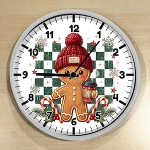 May include: A round wall clock with a white face featuring black numbers and hands. The center displays a gingerbread person wearing a red knit hat with a tag reading MERRY & BRIGHT, holding a coffee cup, set against a green and white checkered background with snowflakes, candy canes, and holly.