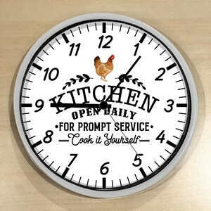 May include: White kitchen clock with black numbers and hands. The clock has a black and white image of a chicken and the text "Kitchen Open Daily - For Prompt Service: Cook it Yourself".
