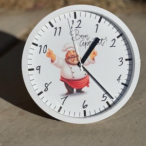 May include: White round clock with black numbers and hands. The clock face features a cartoon chef with the text "Bon Appetit!" in black.
