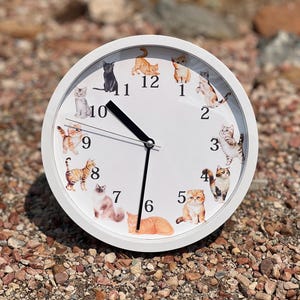 May include: White round clock with black hands and a white face. The numbers 1 through 12 are printed on the face, and each number is surrounded by a different image of a cat.