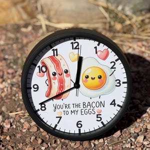 May include: A black and white clock with a cartoon bacon and egg design. The clock face reads "You're the bacon to my eggs" with a small heart on either side of the text.