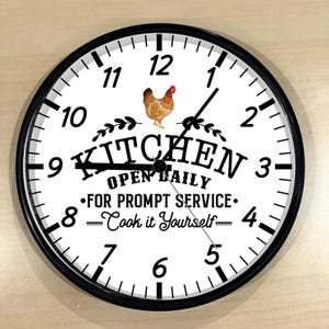 May include: A black and white kitchen clock with a white background and black numbers. The clock has a black border and a black hand. The clock face says "Kitchen Open Daily - For Prompt Service - Cook it Yourself" with a picture of a chicken.