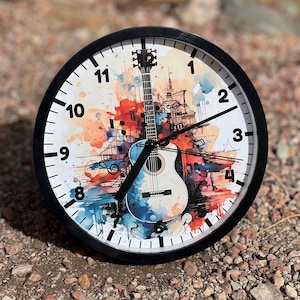 May include: A black and white guitar with watercolor splashes in shades of blue, orange, and red. The guitar is the center of the clock face, with black hands and white numbers. The clock has a black frame.