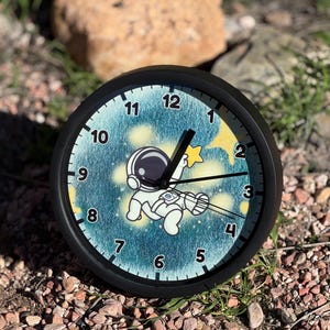 May include: A black-framed clock with a whimsical space theme. The clock face features a cartoon astronaut reaching for a star against a blue and yellow galaxy background. The clock hands are black, and the numbers are white.