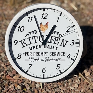 May include: White round clock with black hands and numbers. The clock face has a black and white illustration of a chicken and the words "Kitchen Open Daily" with the text "For Prompt Service - Cook it Yourself -" below.